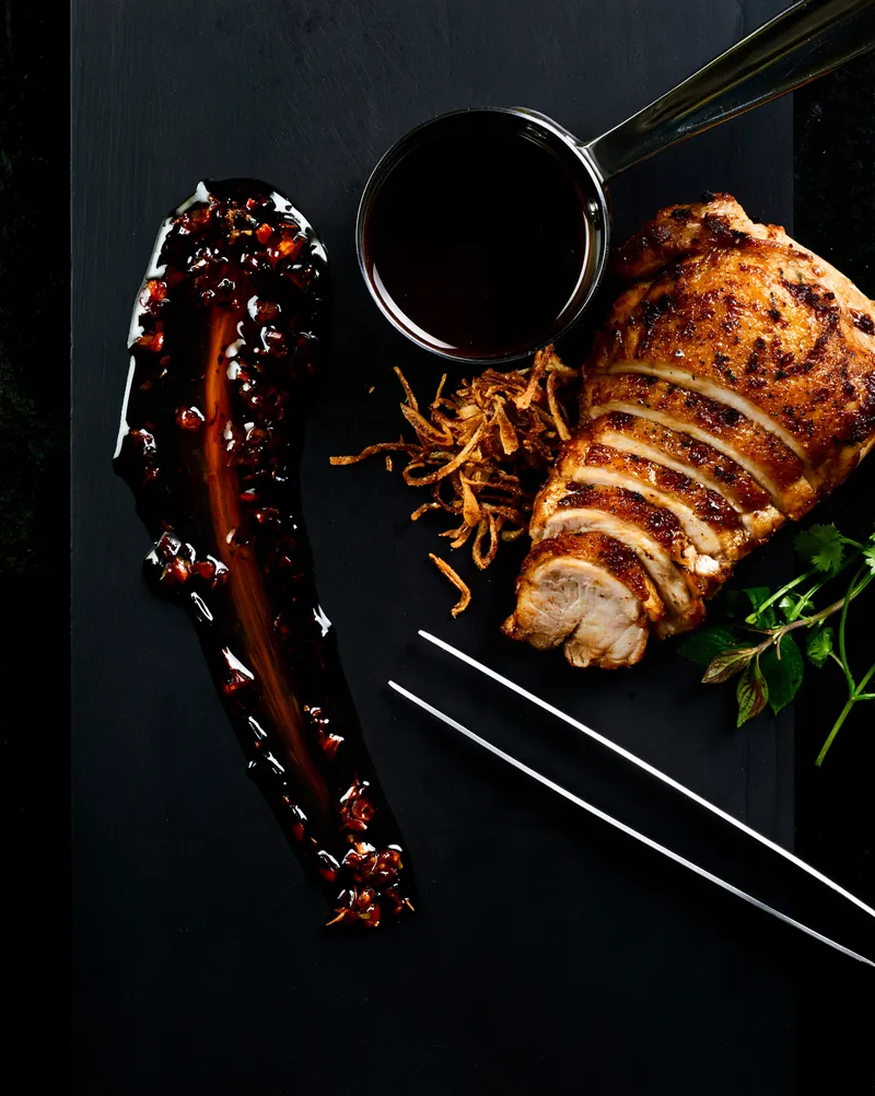A spoon drag of glossy sauce beside seared sliced chicken, crispy shallots, herbs and a small steel saucepan on a matte black surface.